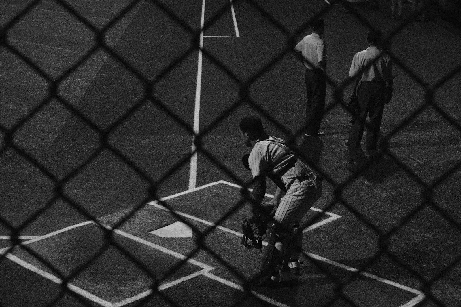 A baseball catcher in crouch seen through a chain-link fence.